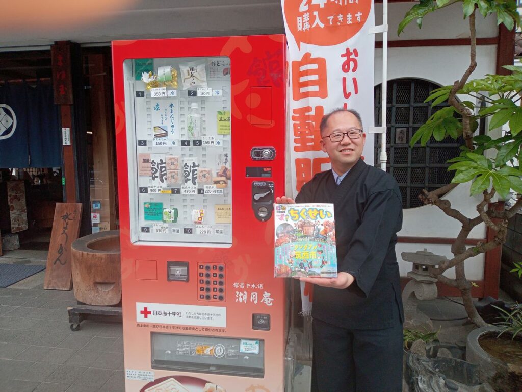 「るるぶちくせい」を手に、自販機の前で筑西市への来訪を呼び掛ける上野さん＝筑西市丙